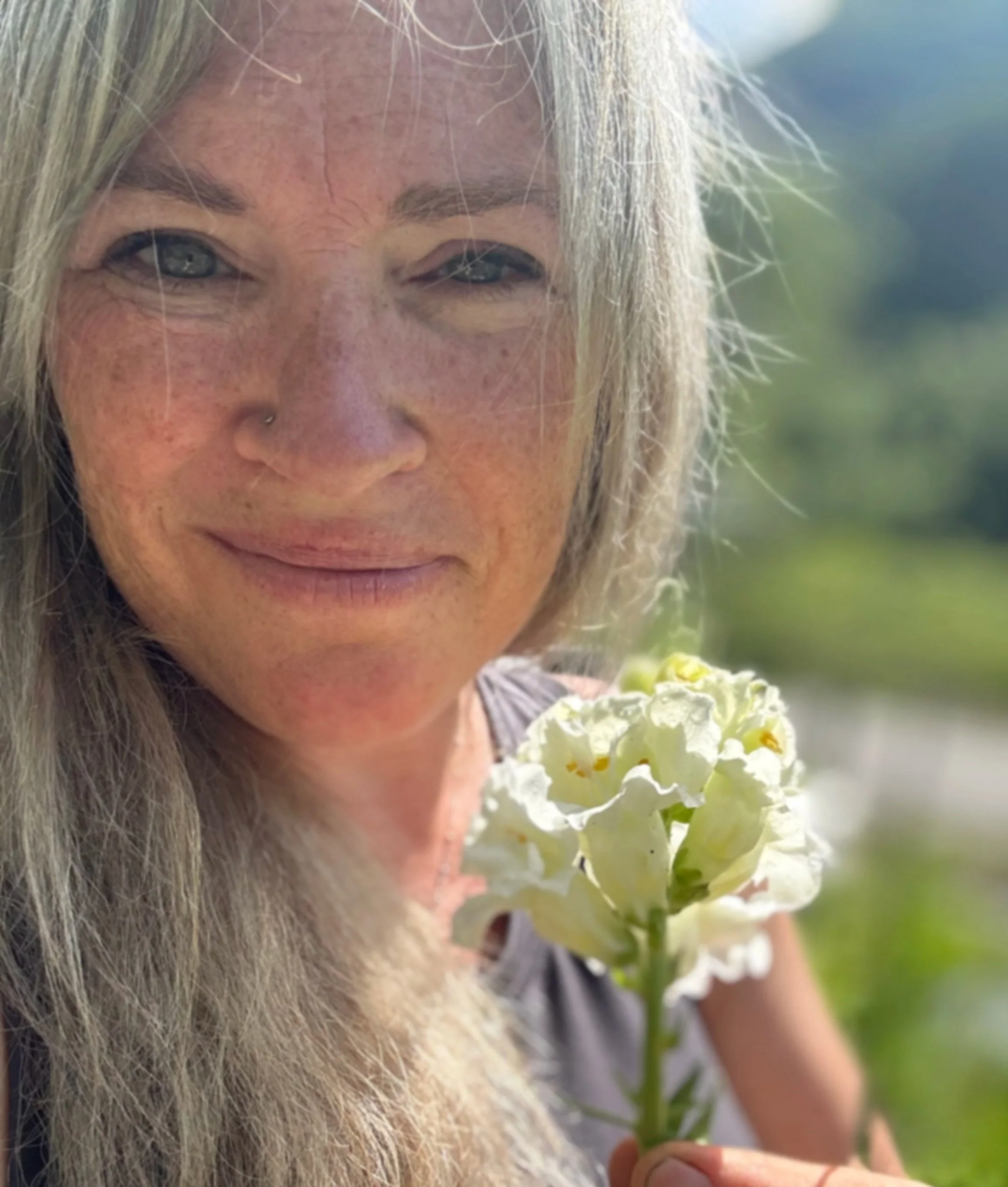 A woman with long blonde hair holding a white flower outdoors, with a blurred green background.