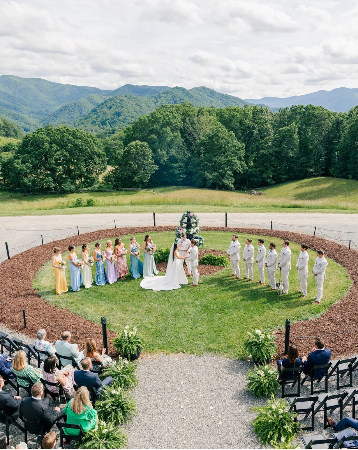 Outdoor wedding ceremony on a grass area with mountains and trees in the background, officiant and bride and groom in the center, bridesmaids in colorful dresses on the left, groomsmen in white suits on the right, seated guests in front.