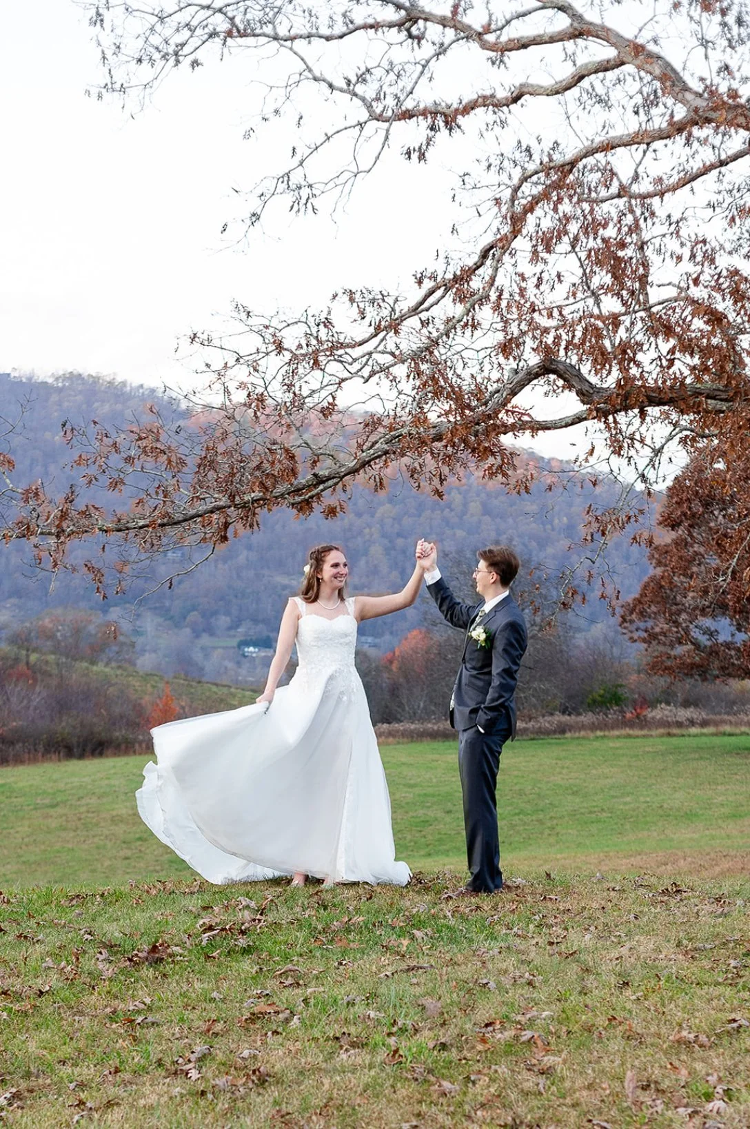 A bride and groom celebrating outdoors on a grassy field, with mountains and autumn trees in the background. The bride is in a white wedding dress, and the groom is in a dark suit. They are holding hands in a victory pose.