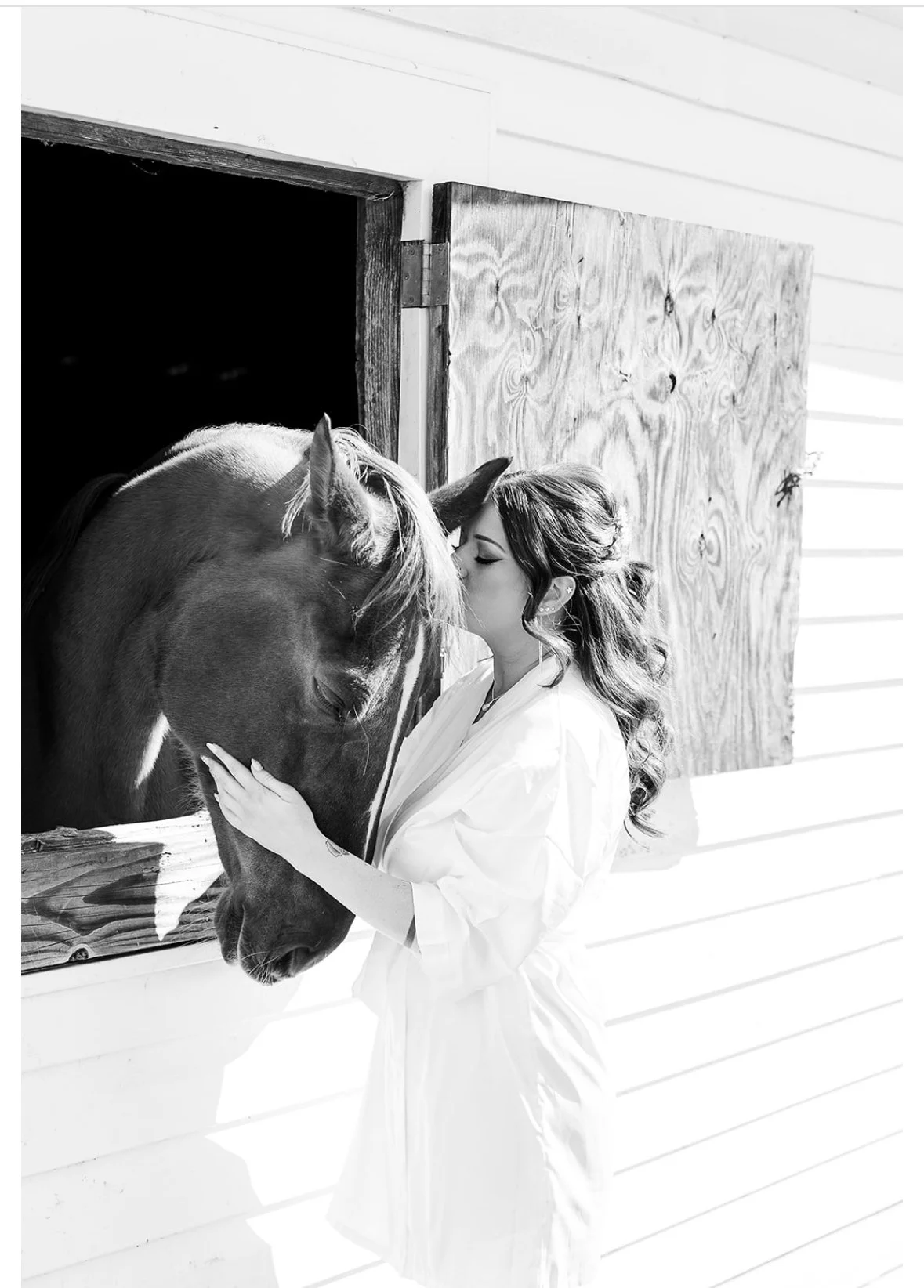 A woman with long wavy hair kissing a horse's face through a barn window, with the barn wall and window opening visible.