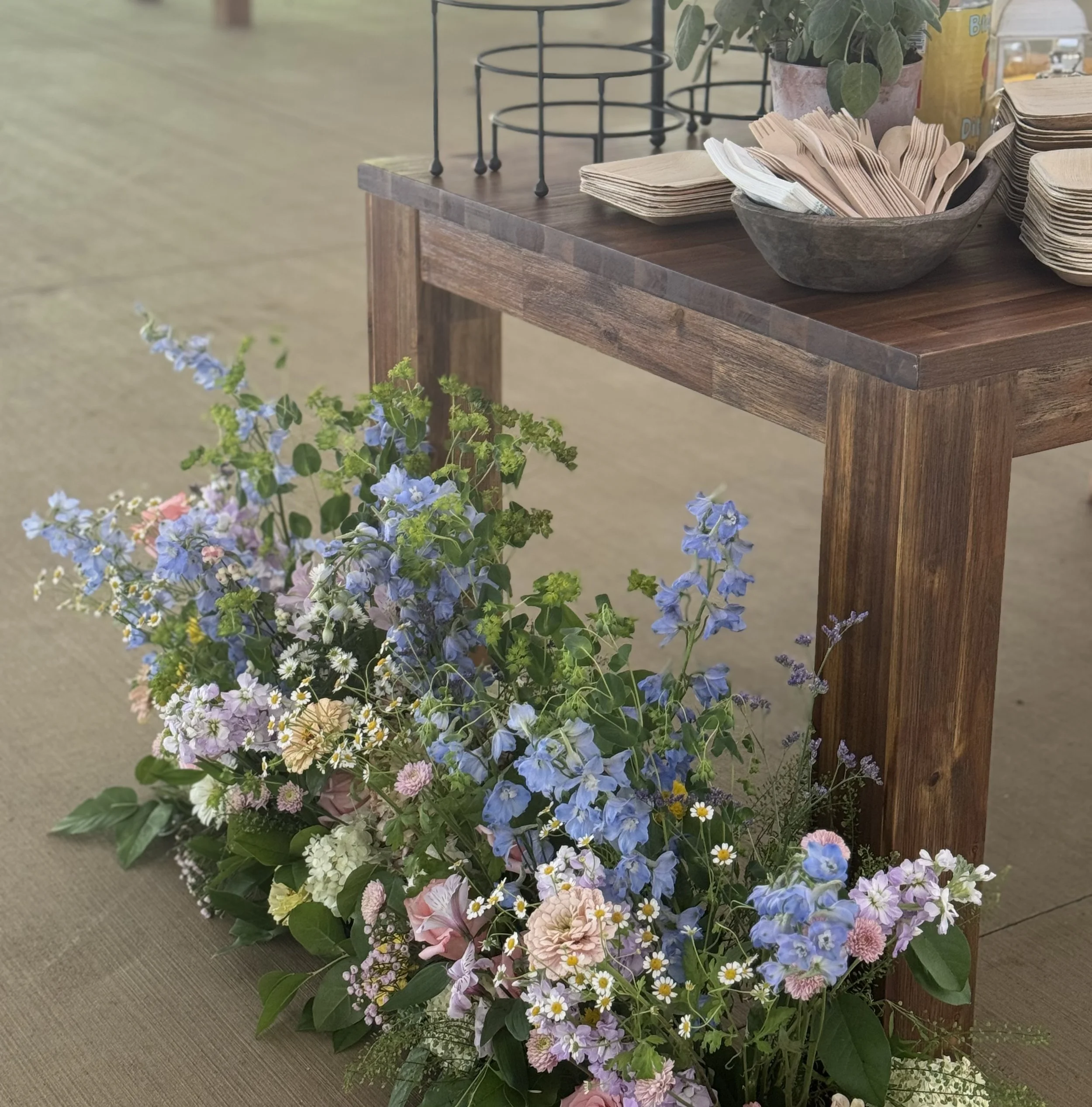 Colorful flower arrangement with blue, pink, purple, and white flowers on the floor next to a wooden table with napkins and plates.