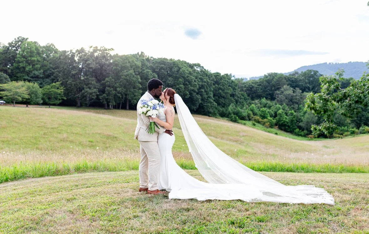 A bride and groom standing close together in a green field, sharing a kiss, with the bride holding a bouquet of flowers and wearing a long white wedding dress with a veil, and the groom in a beige suit.
