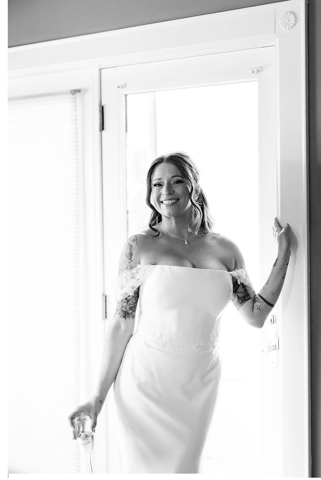 Smiling woman in wedding dress standing by a doorway, holding a glass, with tattoos visible on her arms.
