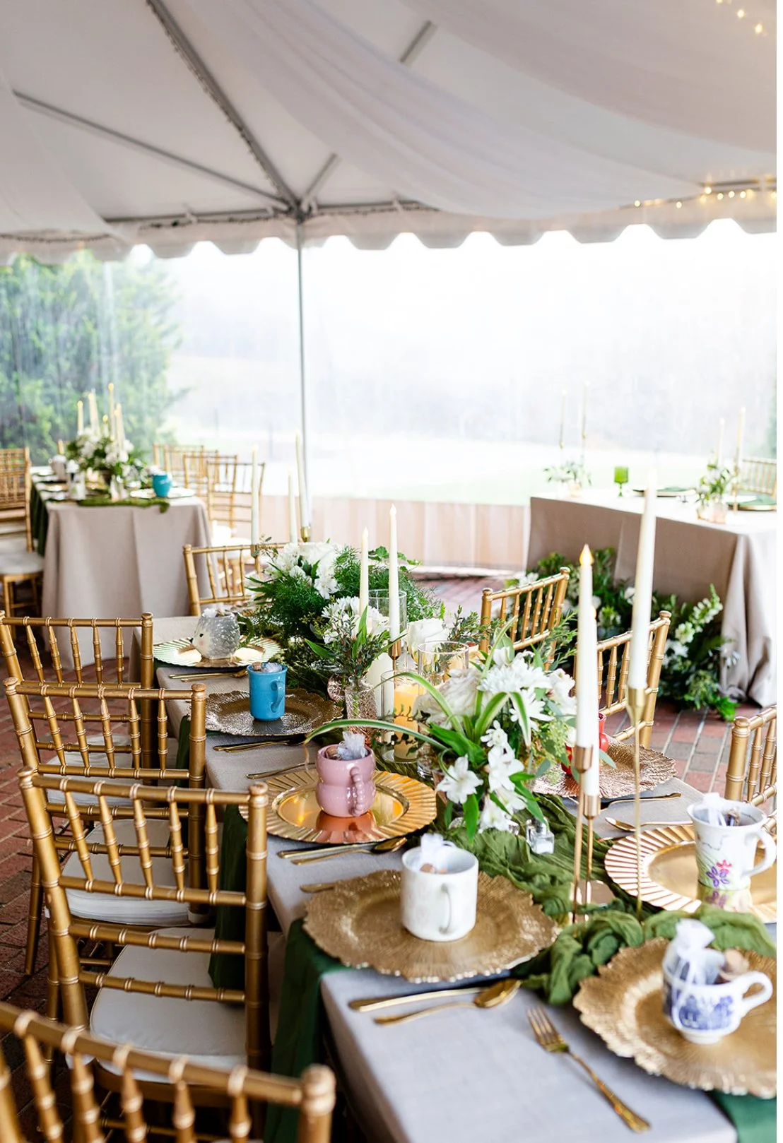 A decorated outdoor wedding or event reception tent with a long table set with gold chargers, floral centerpieces, and assorted cups and mugs, with gold chairs and a backdrop of greenery and a lake.