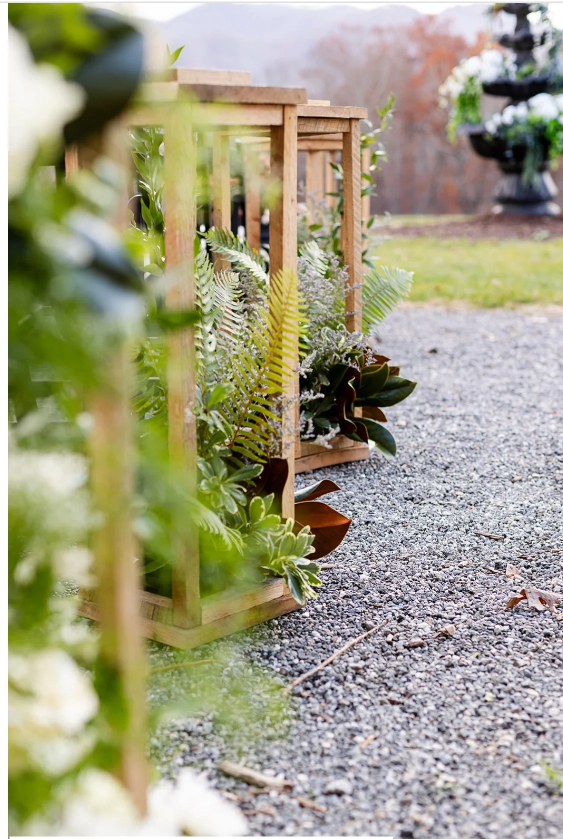 Wooden planter boxes filled with various green plants and ferns, placed outdoors on a gravel path with a grassy area and decorative urn in the background.