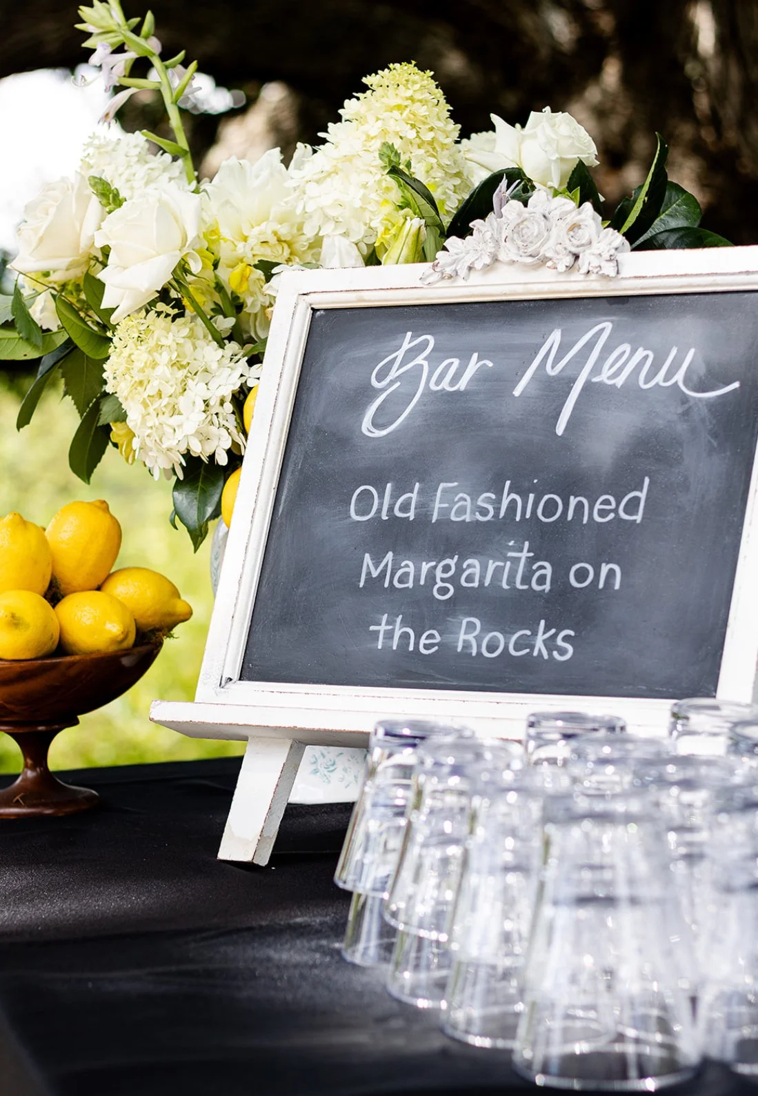 A chalkboard sign with white cursive lettering that reads 'Bar Menu: Old Fashioned Margarita on the Rocks,' surrounded by a flower arrangement of white roses, hydrangeas, and greenery, with lemons in a bowl and empty glasses in front, outdoors settin