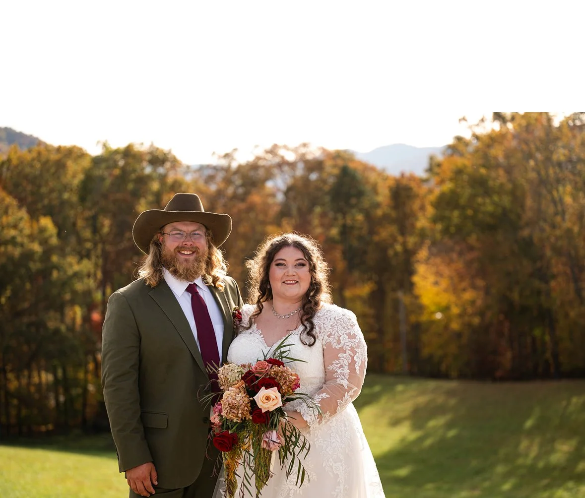 A smiling bride and groom standing outdoors with autumn trees in the background. The bride is holding a bouquet of flowers and wearing a white lace wedding dress. The groom is wearing a green suit, a white shirt, a burgundy tie, and a wide-brimmed ha