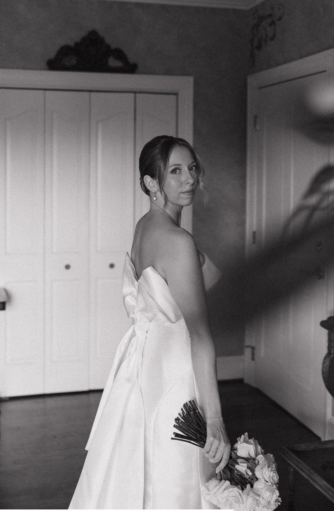 A woman in a wedding dress holding a bouquet of roses, standing indoors in front of a wardrobe, looking at the camera.