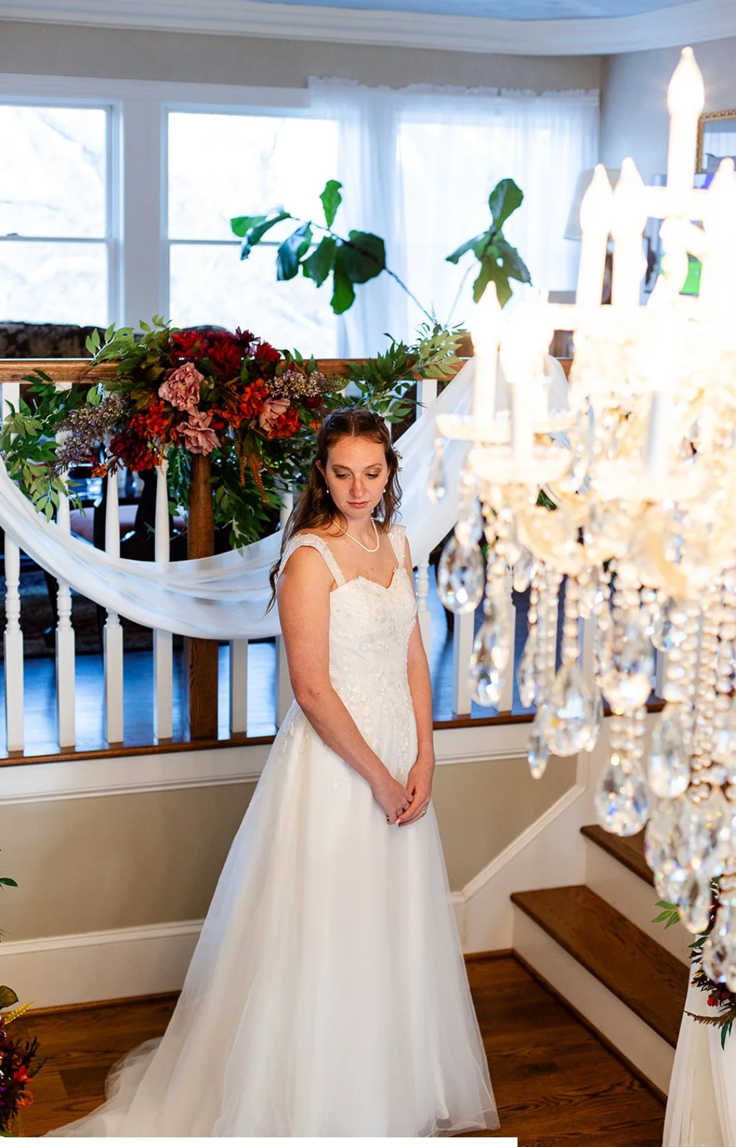 A bride in a white wedding dress standing on a staircase, looking down with her hands clasped in front of her. There is a chandelier hanging in the foreground and a floral arrangement with greenery behind her, near large windows with white curtains.