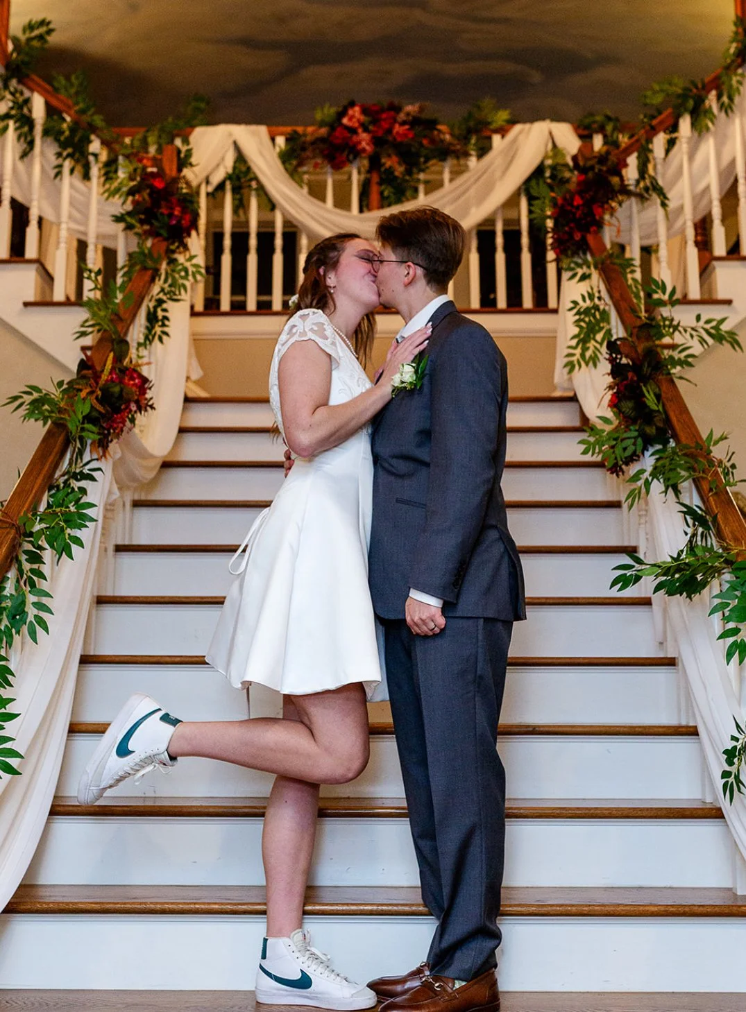 A couple kissing on a staircase at their wedding, decorated with greenery and flowers.