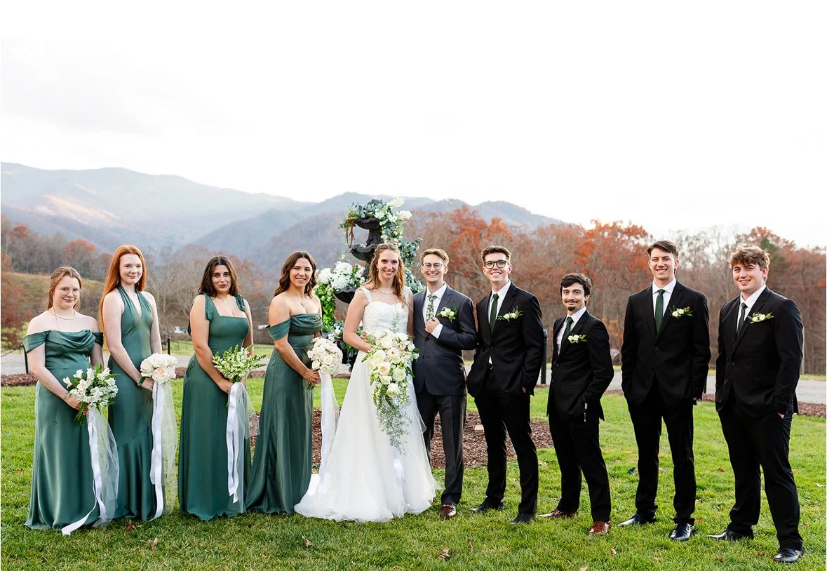 A group of ten people, including a bride and groom, standing outdoors on grass with mountains and autumn trees in the background. The women are wearing green bridesmaid dresses and holding white bouquets, while the men are in black suits with white s