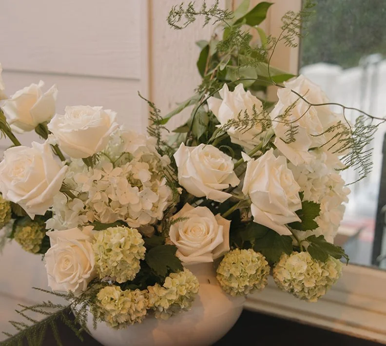 A white vase filled with white roses, hydrangeas, and greenery placed on a windowsill.