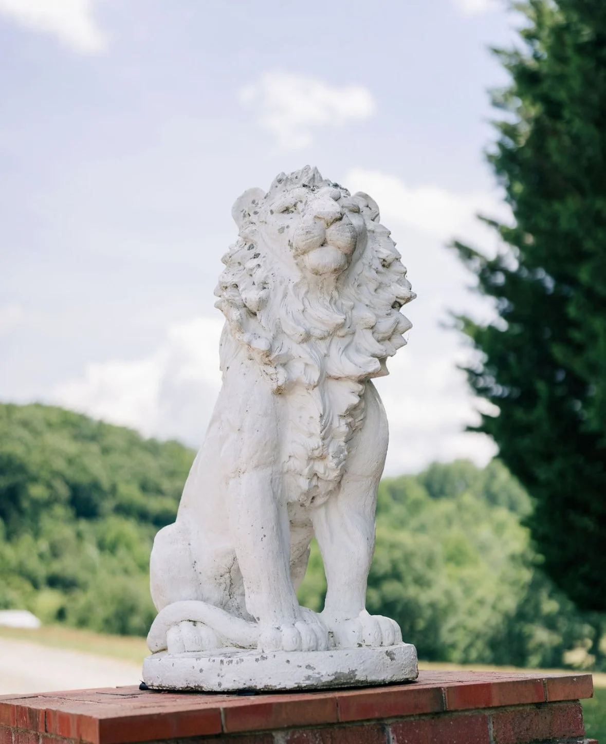 White stone lion sculpture on a brick pedestal outdoors, with green trees and a partly cloudy sky in the background.