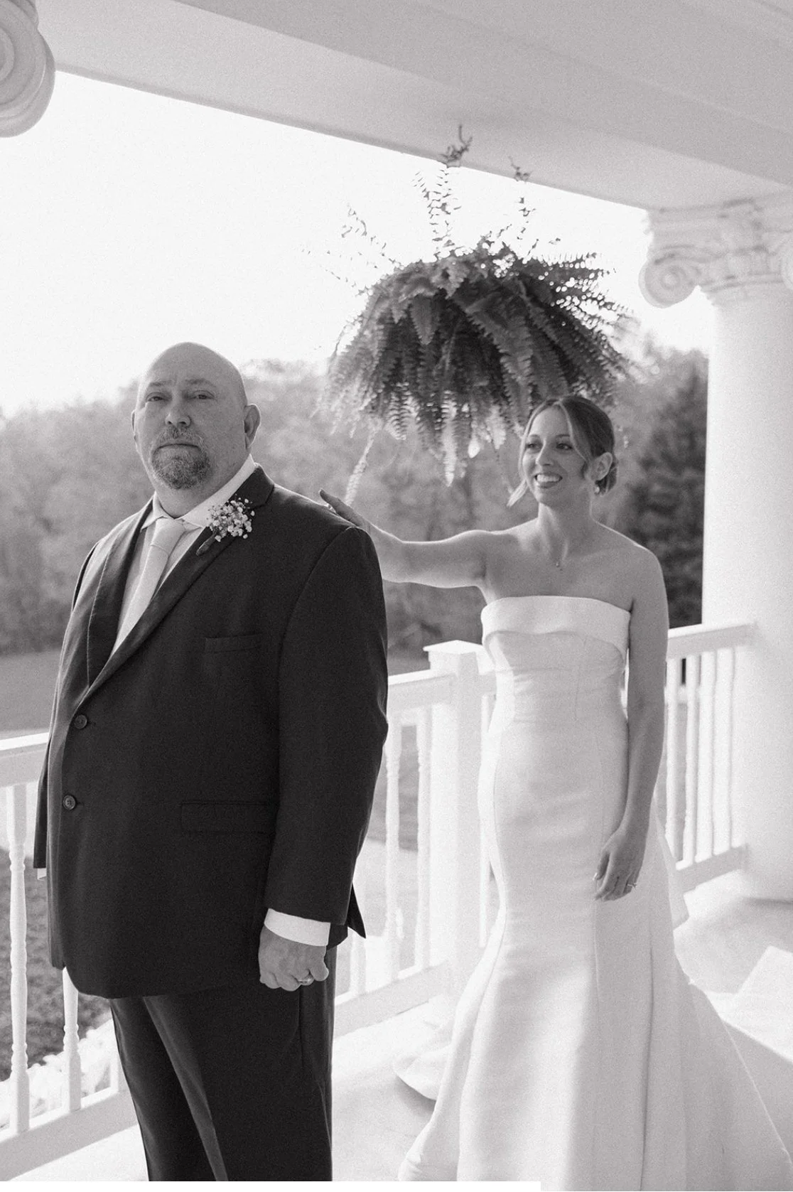 A woman in a strapless wedding gown smiling and gently touching the shoulder of a man in a suit, standing on a porch with a white railing and a large fern hanging in the background.