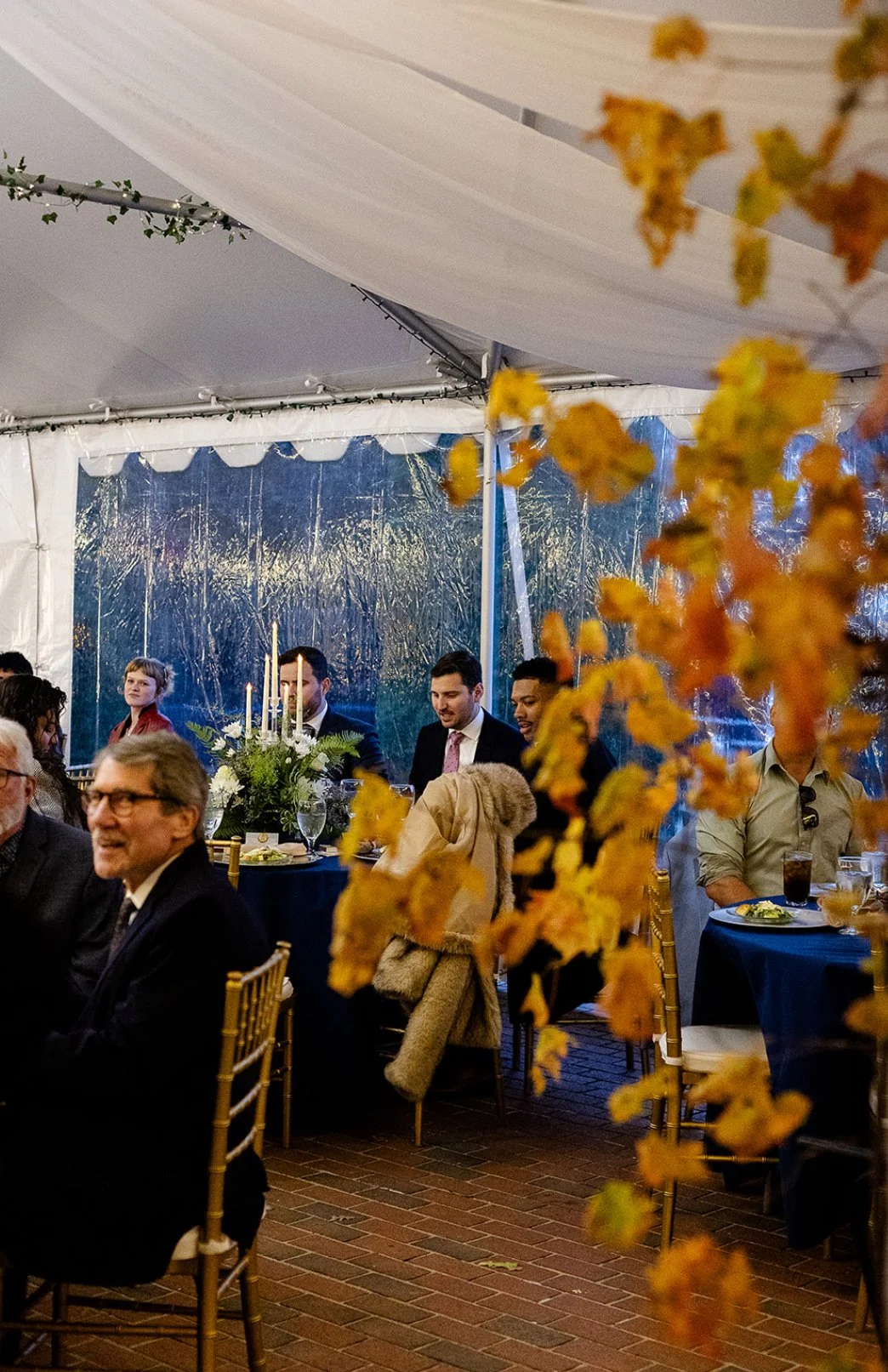 People attending a formal event or dinner inside a decorated tent, with tables set with candles, flowers, and glassware, and autumn leaves partially obscuring the view.