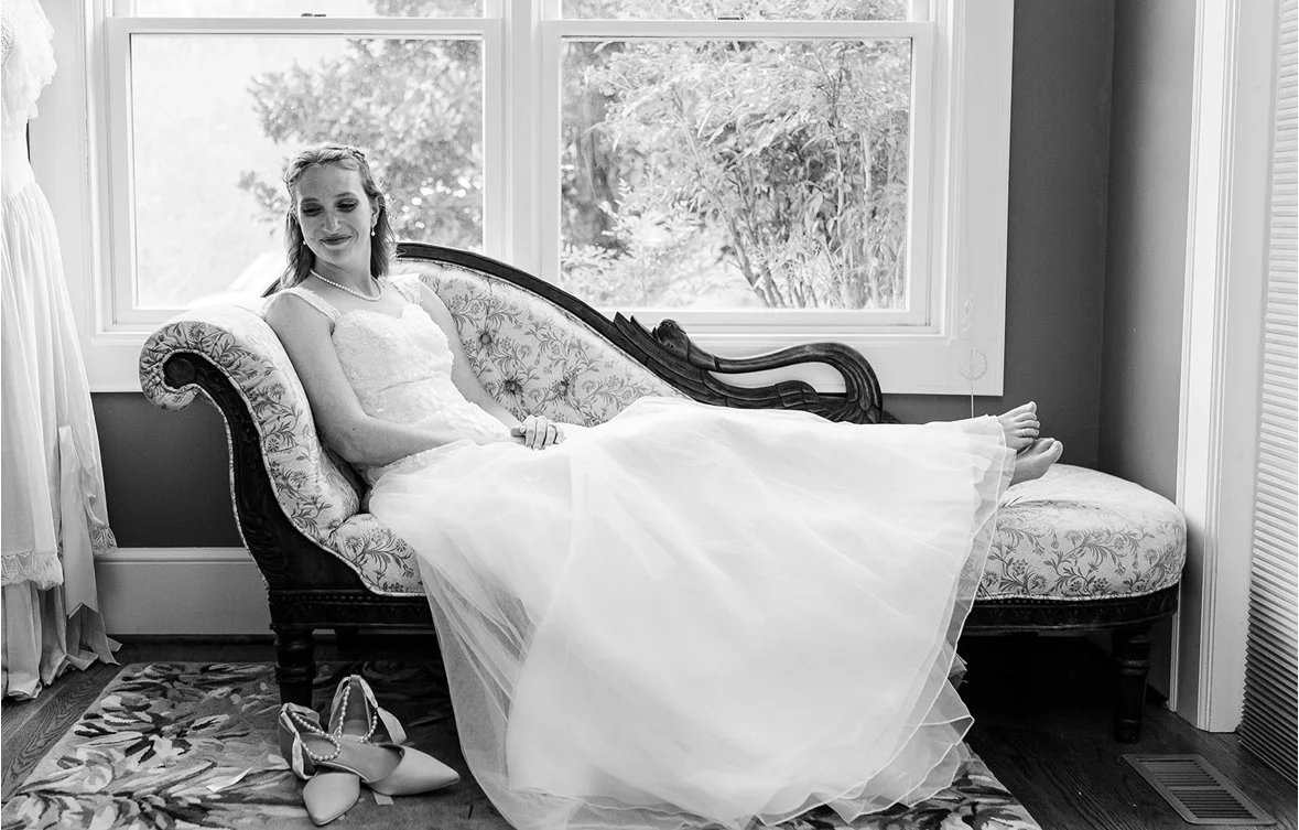 A bride in a wedding dress sitting on a vintage upholstered settee in front of large windows, with her shoes on the floor nearby.