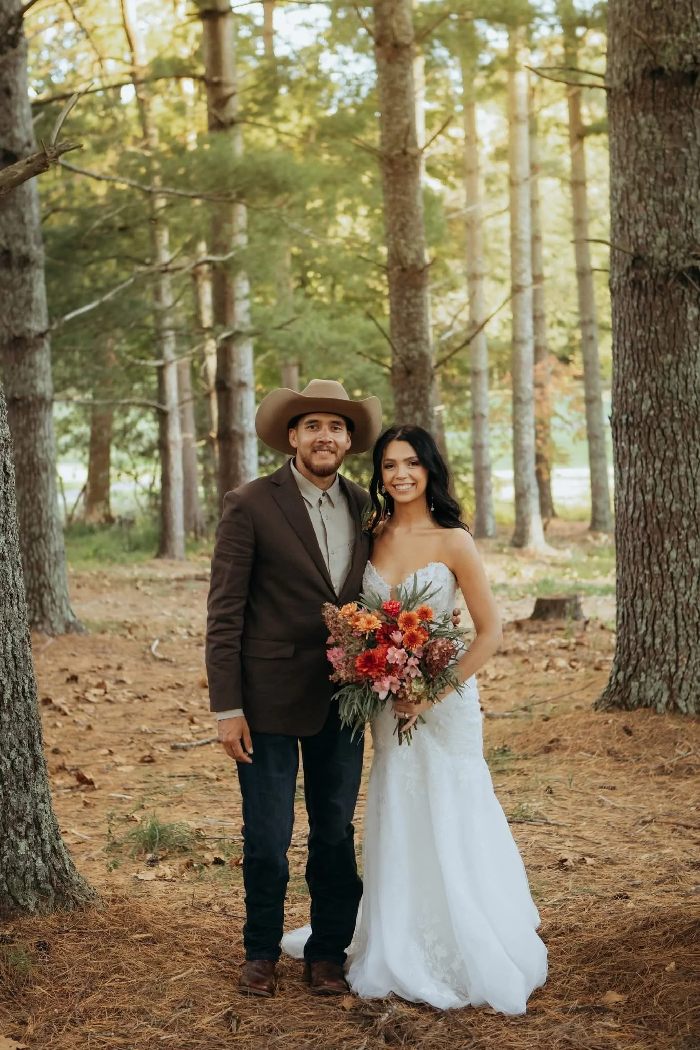 A newlywed couple standing in a wooded forest during daytime. The groom is wearing a brown suit with a light-colored shirt and a large cowboy hat. The bride is dressed in a strapless white wedding gown, holding a bouquet of colorful flowers, and has 