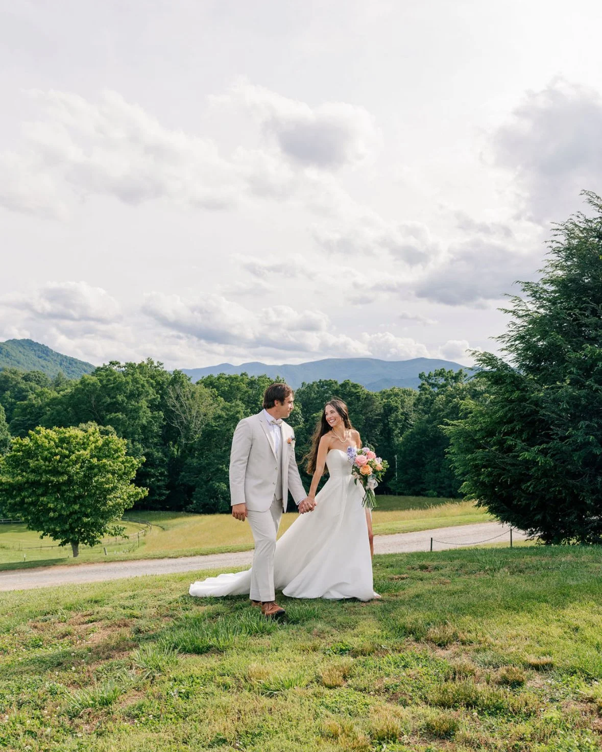 A bride and groom holding hands in a scenic outdoor setting with green trees and mountains in the background, during daytime.