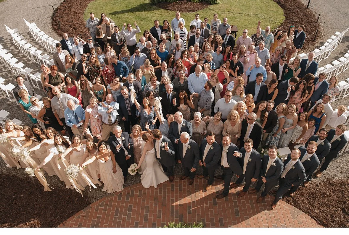 Group photo of wedding guests outdoors, with the bride and groom at the front center, surrounded by family and friends, all facing the camera.