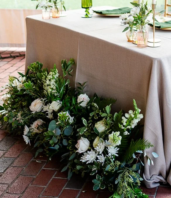 A wedding or event table decorated with a beige tablecloth, elegant green glassware, white plates, and small flower arrangements, with a large white floral arrangement and greenery on the floor beside the table on a brick patio.