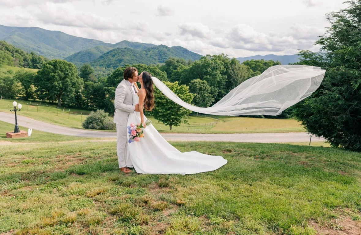 A bride and groom share a kiss outdoors on a grassy hill, with the bride holding a bouquet, in a scenic landscape with mountains and trees in the background.