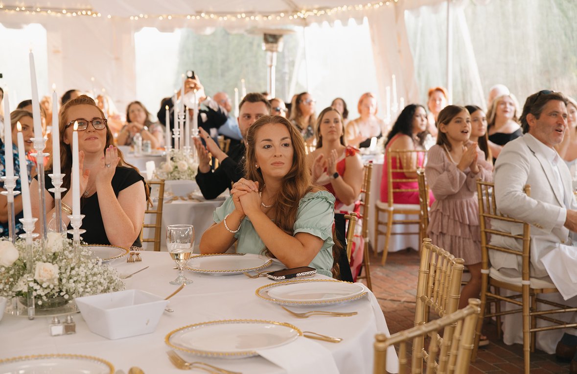 Guests seated at decorated tables during a wedding reception, clapping and watching a performance or speech, under a tent with string lights.