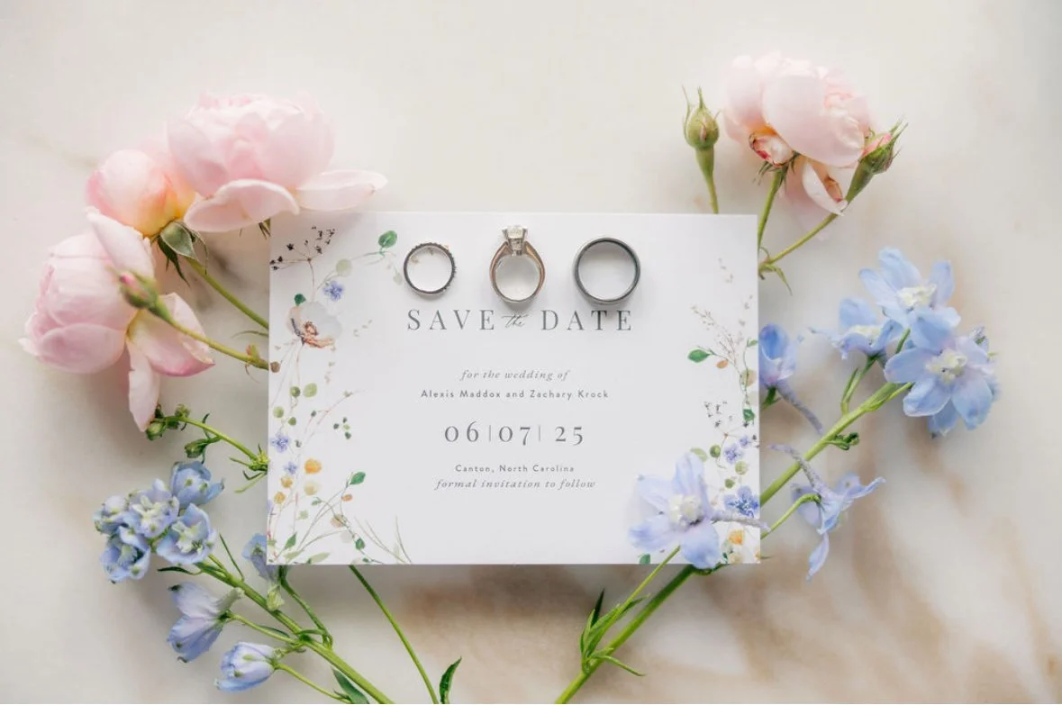 Save the date card for a wedding, surrounded by pink and light blue flowers, with three rings placed on the card.