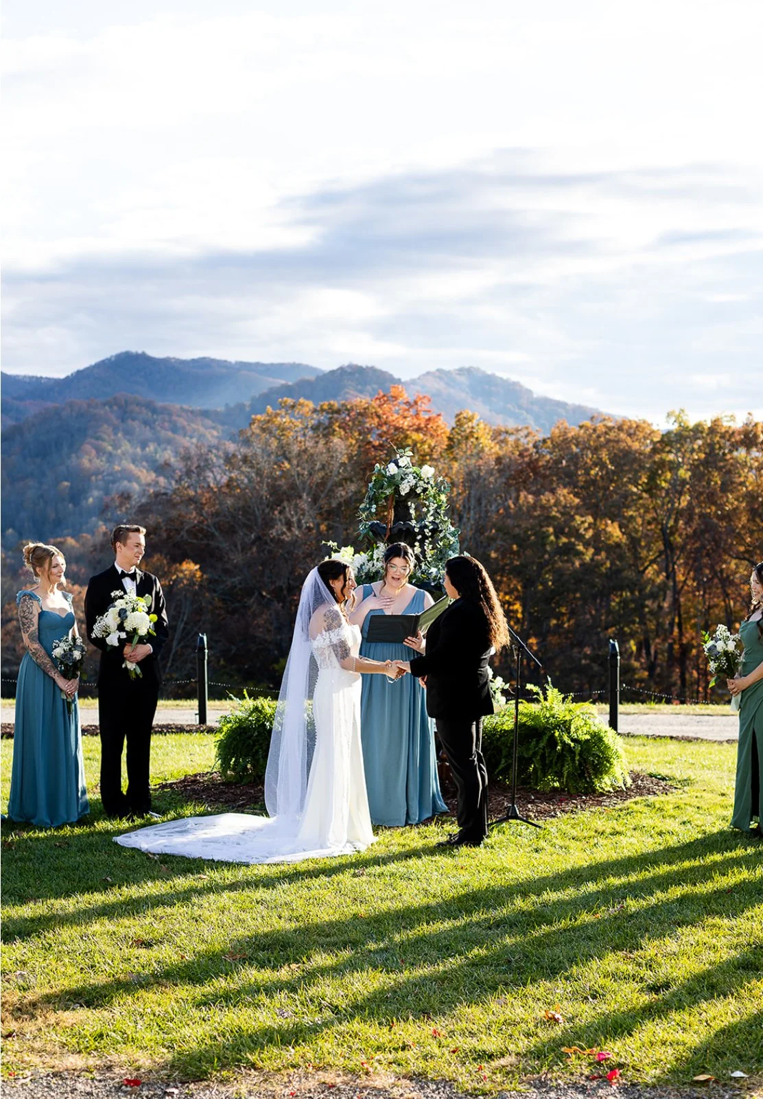A wedding ceremony outdoors with mountains and autumn trees in the background, where a bride and groom are exchanging vows, surrounded by bridesmaids and groomsmen.