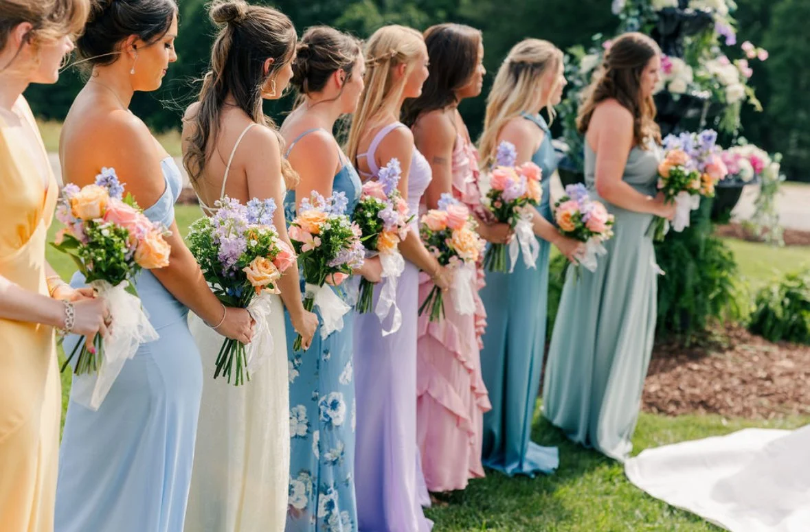 Group of women in colorful dresses holding bouquets of flowers at an outdoor wedding ceremony.