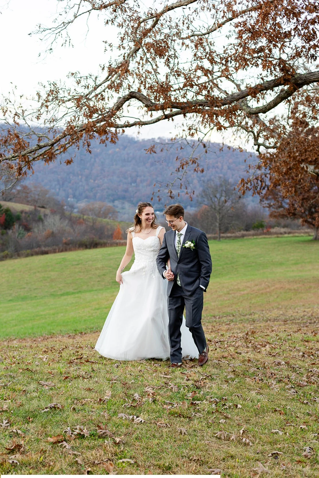 A bride and groom walking together outdoors on a grassy field, smiling and holding hands, with a scenic background of trees and mountains during fall.