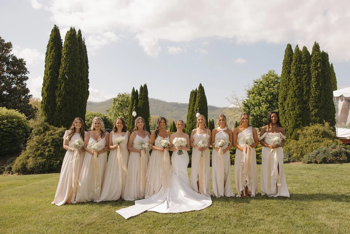 Bride and her nine bridesmaids standing outdoors on a sunny day with green trees and hills in the background, all wearing cream-colored dresses and holding bouquets.