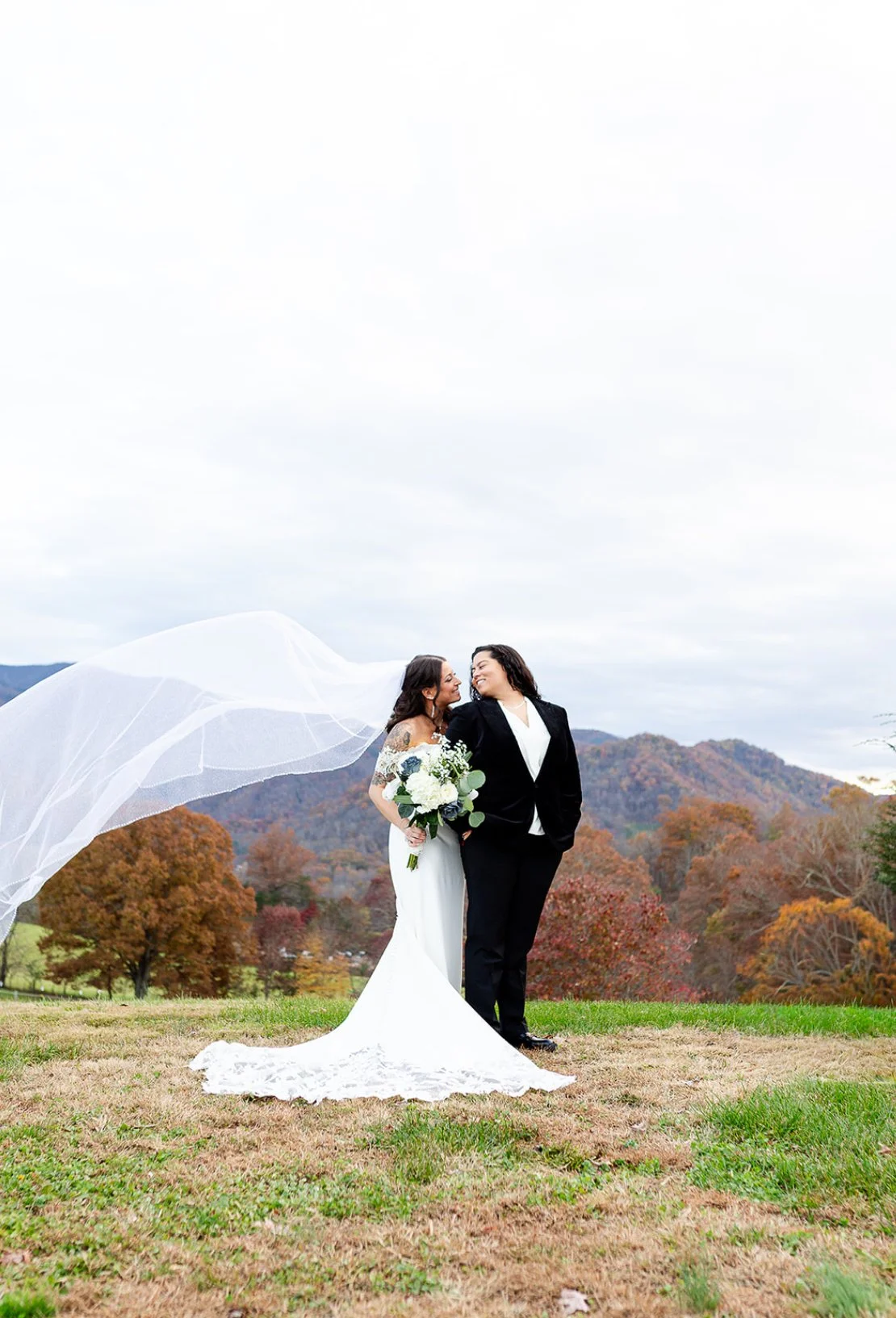 Two women, one in a white wedding dress and the other in a black suit, standing close and smiling at each other outdoors with a backdrop of trees with fall foliage and mountains.