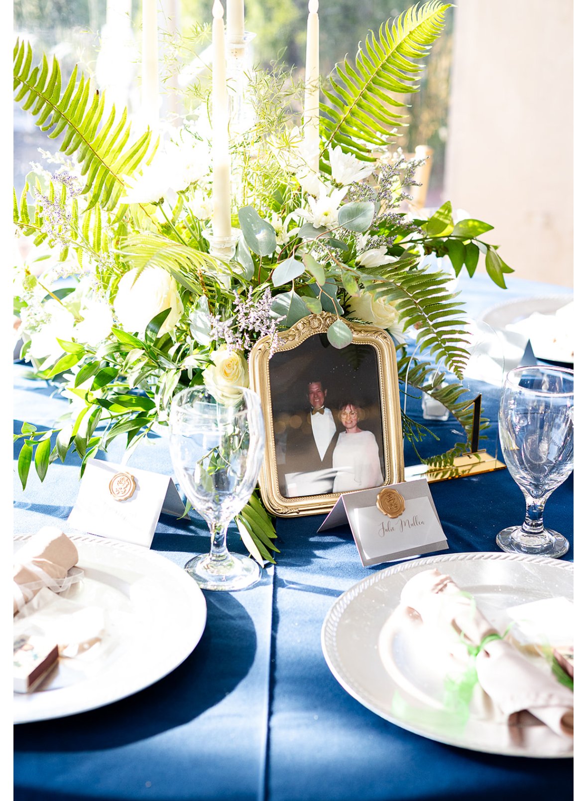 A table arrangement with a gold framed photograph of a couple, surrounded by a lush floral centerpiece with white flowers and greenery, and place cards with gold seals, set for a wedding reception.