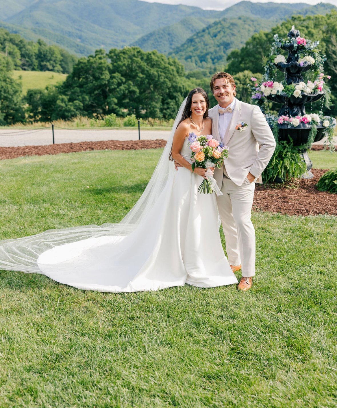 A bride and groom smiling and standing close together outdoors on a grassy field with mountains and trees in the background. The bride is holding a bouquet of flowers, and the groom has his arm around her.