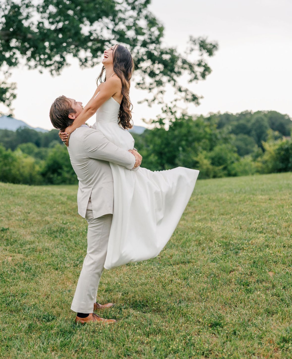 A man in a beige suit lifts a woman in a white wedding dress in a grassy outdoor setting with trees and hills in the background, both laughing.