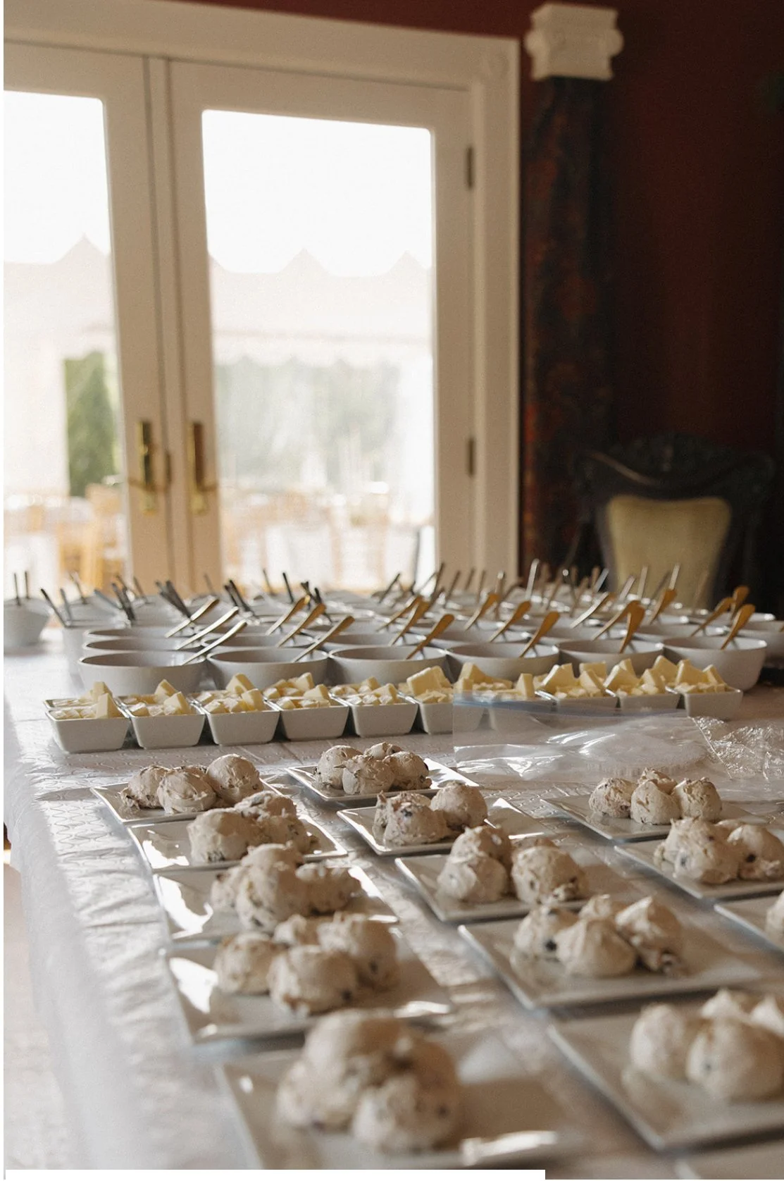 A table with plates of scones, bowls of butter, and small bowls with spoons, set up for a gathering or event, with a view of a bright outdoor area through glass double doors in the background.
