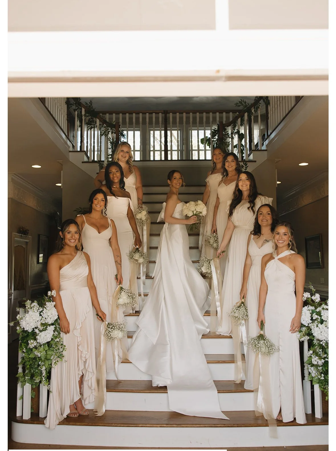 Bride with ten bridesmaids on a staircase indoors, all wearing white dresses, some holding bouquets, with floral decorations on the sides.
