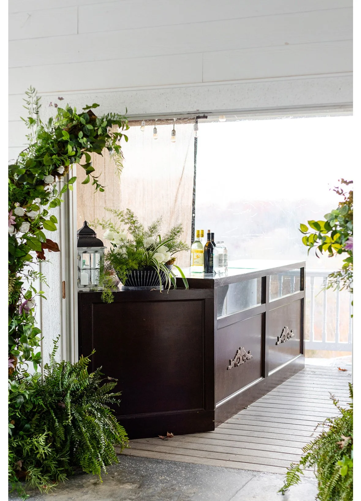 An outdoor bar with a dark wooden counter, decorative handles, and glass panels on the side, decorated with plants and bottles of alcohol, surrounded by lush greenery and plants on the sides.