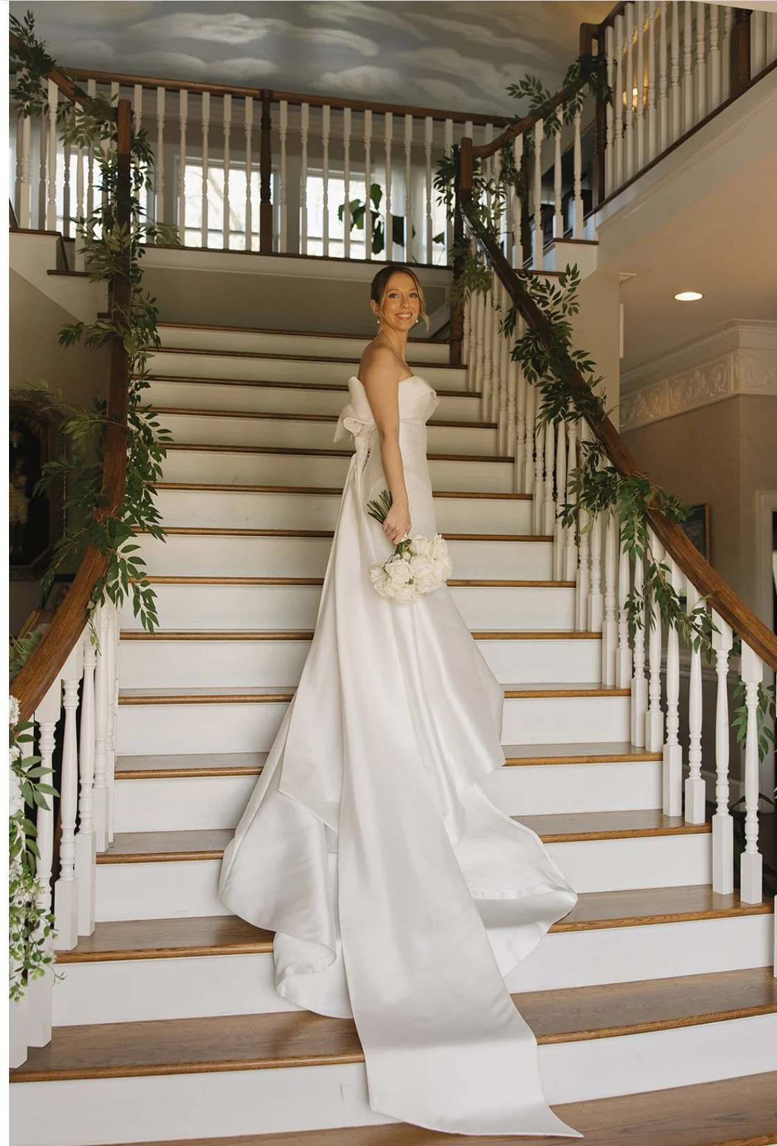 A bride in a white wedding gown holding a bouquet of white flowers standing on a staircase indoors.