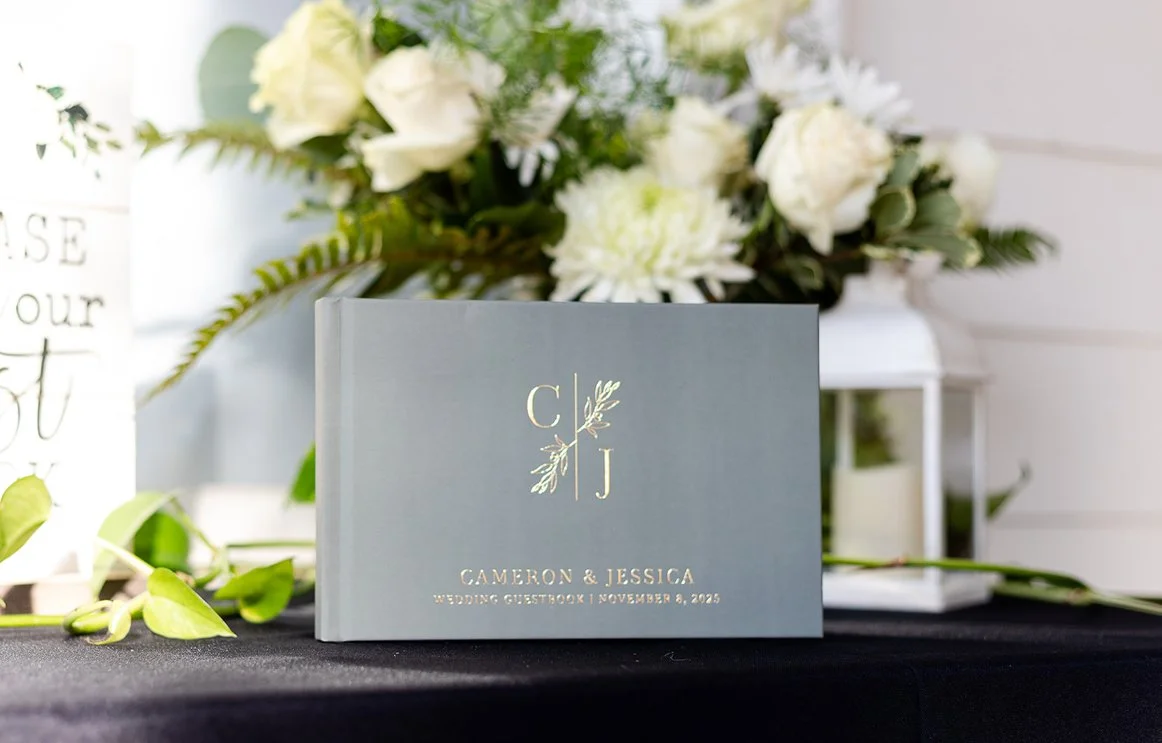 Wedding guestbook titled "Cameron & Jessica" with a monogram "C J" and the date November 8, 2025, placed on a table with floral arrangements in the background.