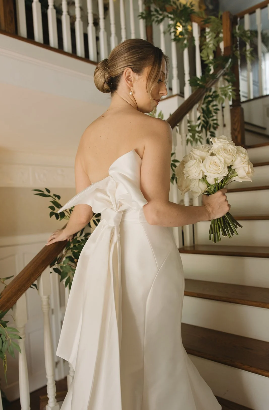 A bride in a strapless wedding gown holding a bouquet of white roses on a staircase with wooden handrail and greenery.