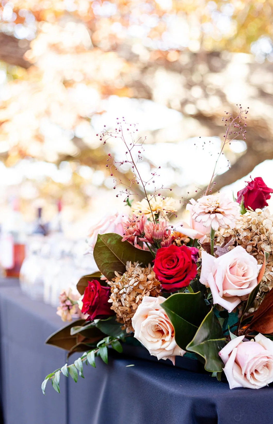 A floral arrangement with red and pale pink roses, beige hydrangeas, and various greenery on a dark tablecloth with a blurred outdoor background.
