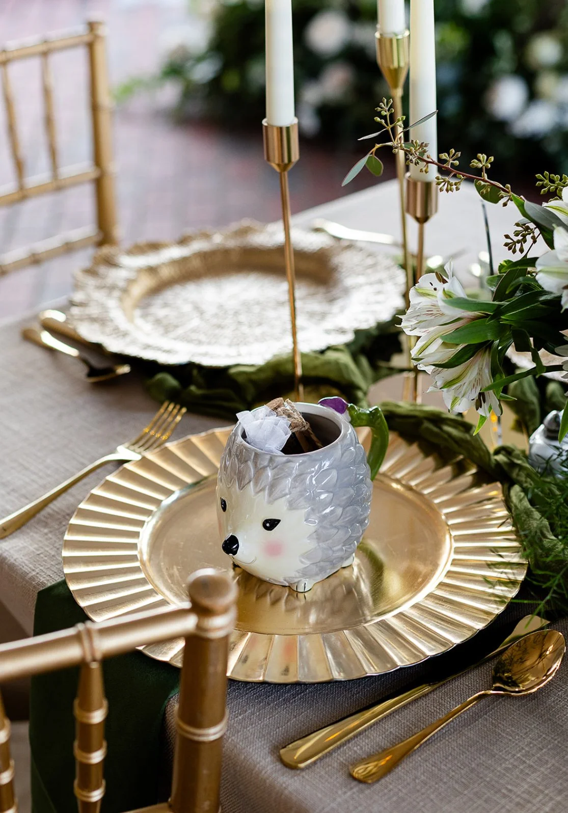 Table setting with a gold charger plate, gold utensils, a hedgehog-shaped mug filled with cinnamon sticks and napkins, floral arrangements, and candleholders.