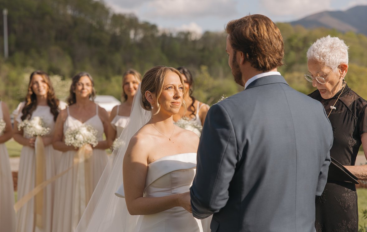 A bride and groom exchanging vows during an outdoor wedding ceremony, with bridesmaids in the background holding bouquets, and an officiant reading from a book, set against a scenic landscape with hills and trees.