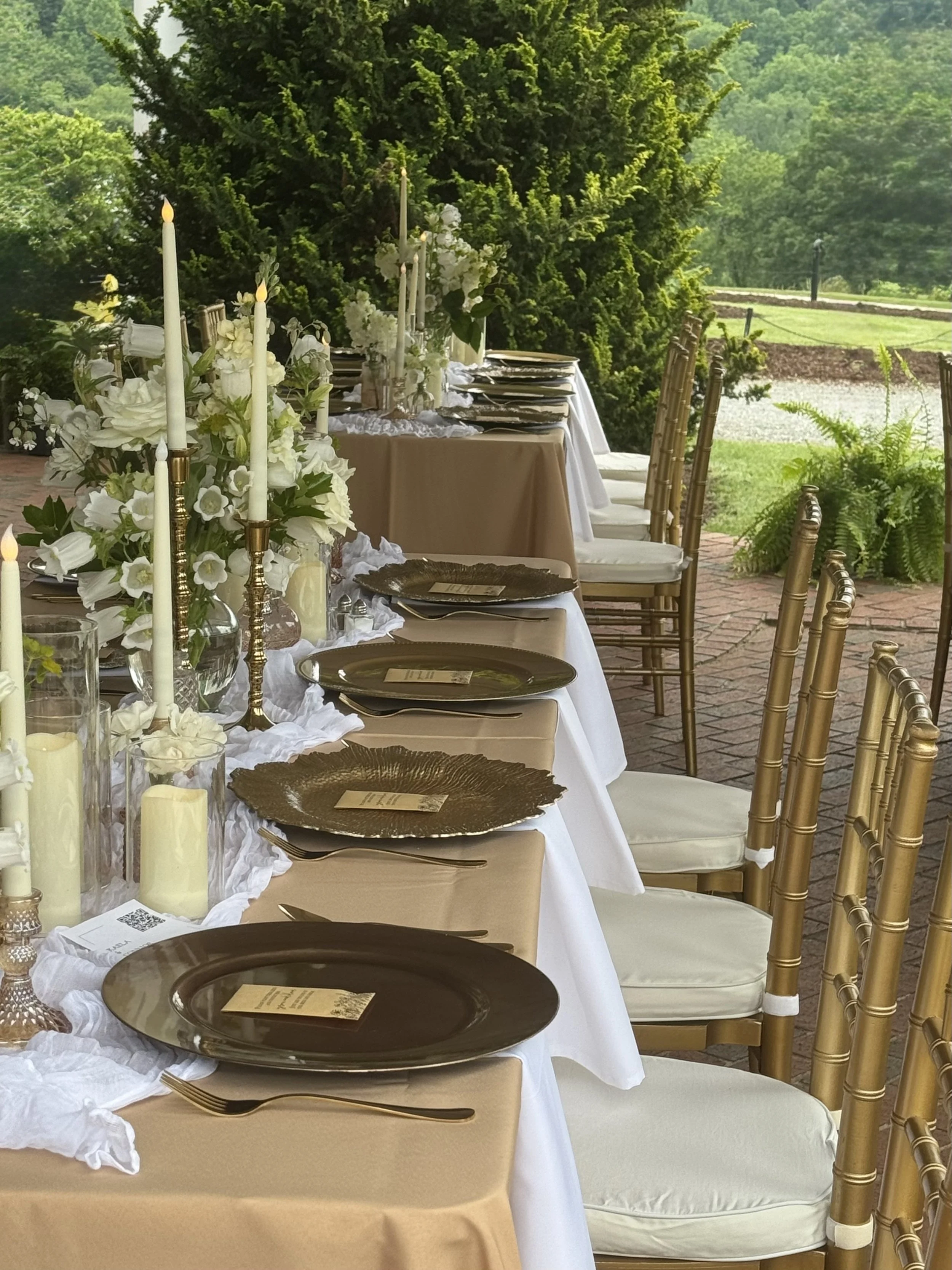 Elegant outdoor dining table set for a celebration, decorated with white flowers, tall white candles, gold flatware, and gold chargers, with gold chairs on a brick patio surrounded by greenery.