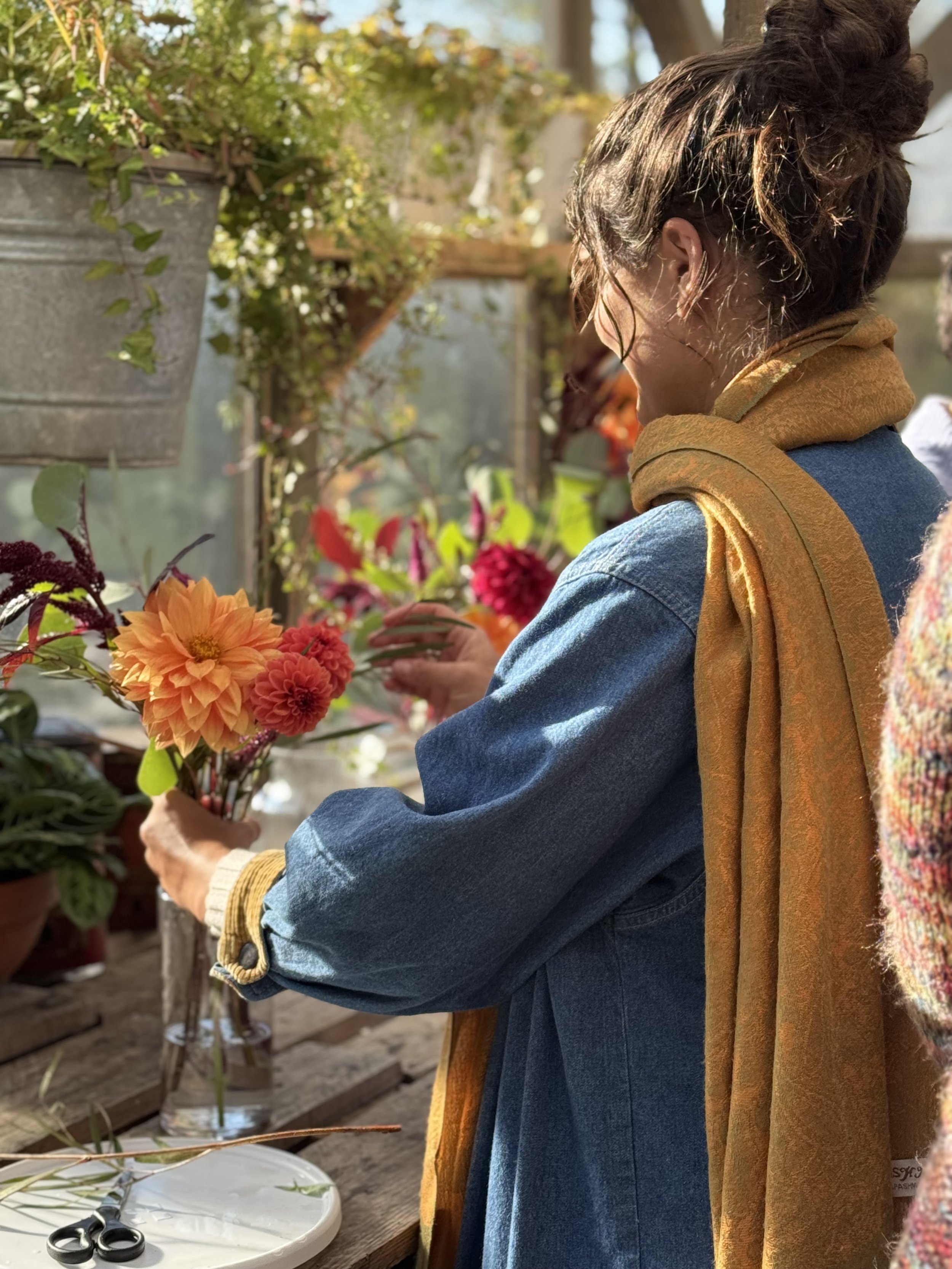 A woman arranging pink and orange flowers in a glass vase, standing in a greenhouse or garden center with sunlight filtering through the windows.