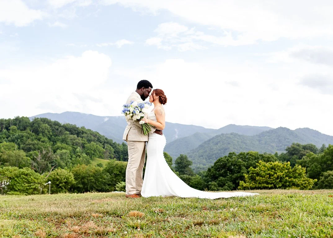 A bride and groom stand close together on a grassy field with rolling hills and trees in the background, outdoors on a cloudy day.