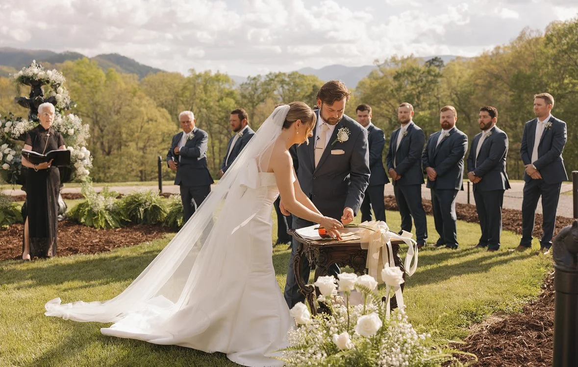 A bride and groom exchange rings during their outdoor wedding ceremony. The bride is in a white wedding dress with a long veil, and the groom is in a dark suit. Bridesmaids and groomsmen stand behind them, with a officiant reading nearby. The setting