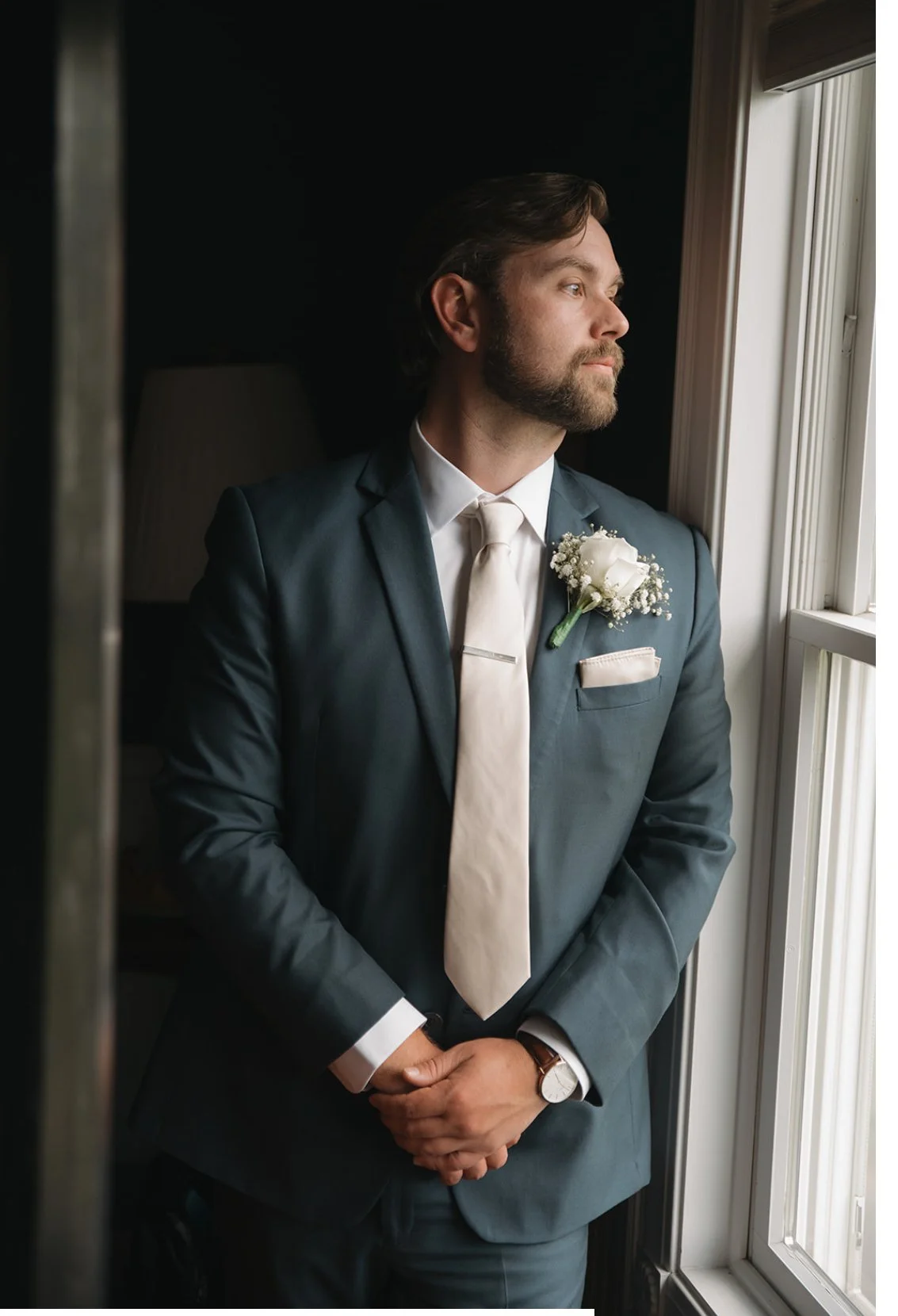 A man in a blue suit with a white shirt and tie, looking out the window, standing in a dimly lit room with sunlight coming in.