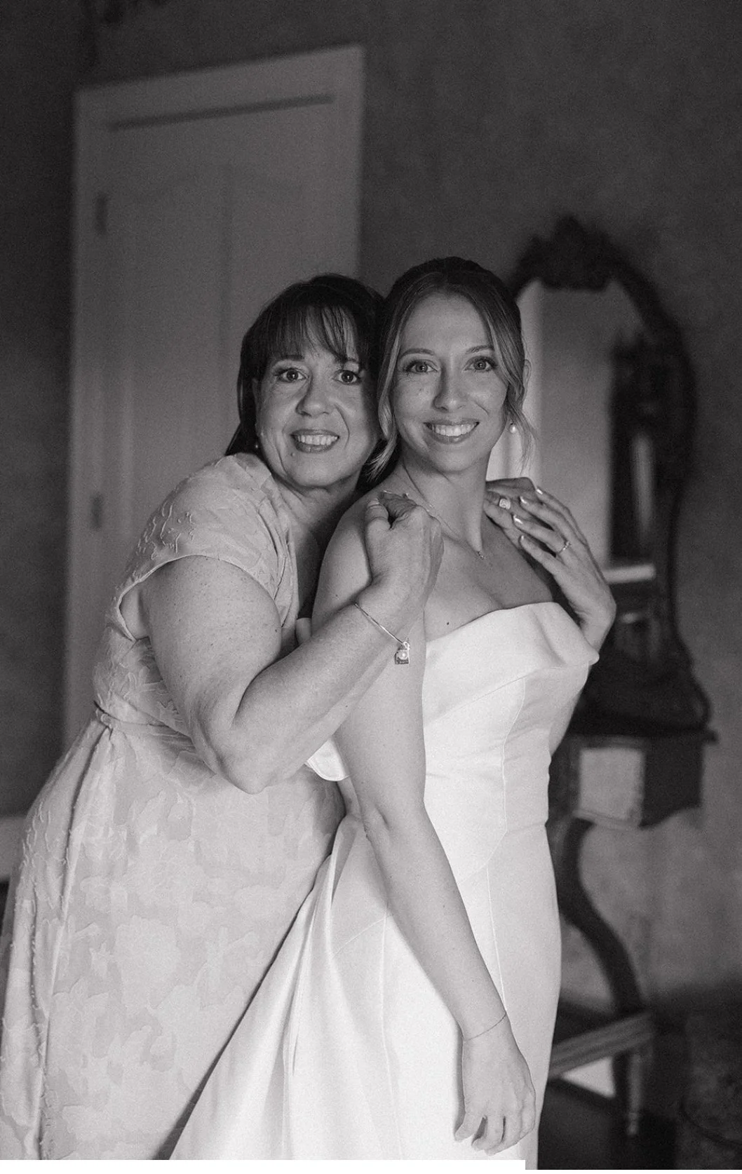 A black and white photo of two women, one younger and one older, smiling and posing together. The younger woman is dressed in a wedding gown, and the older woman is wearing a light-colored dress. They are standing close, with the older woman holding 